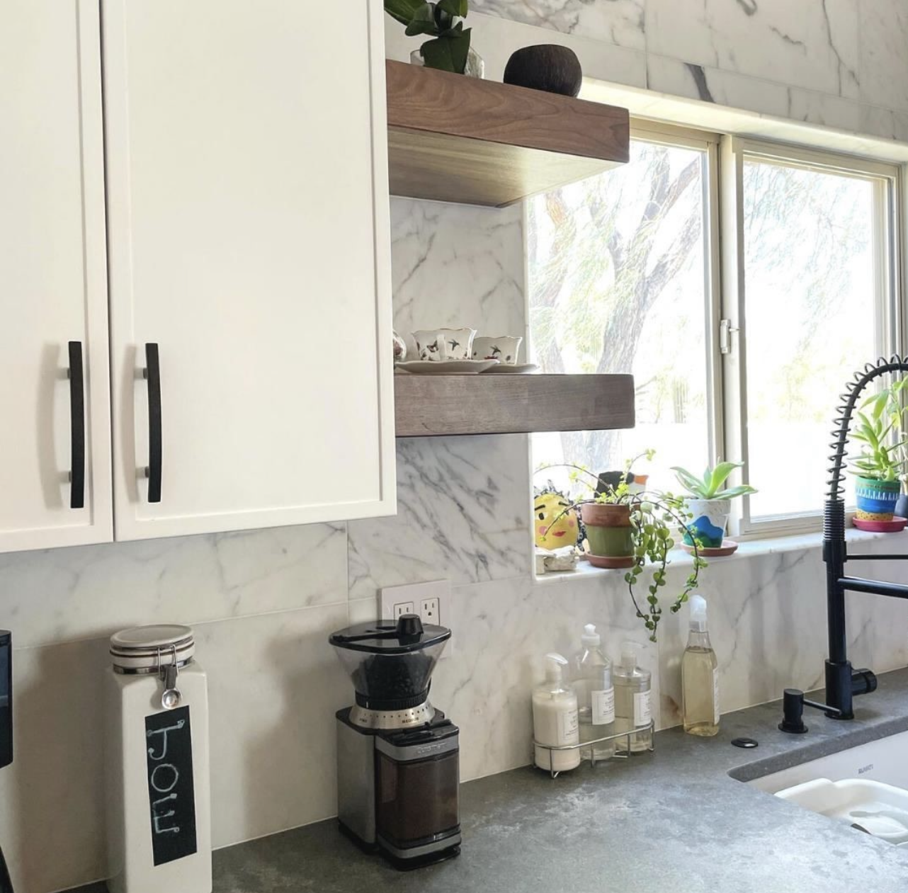 Bright kitchen with white cabinets, marble backsplash, wooden shelves, coffee grinder, sink, plants, and a sunlit window