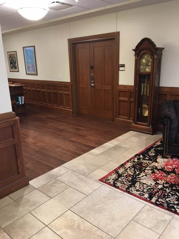Traditional interior hallway with wood-paneled walls, double wooden doors, grandfather clock, tiled floor, and patterned area rug