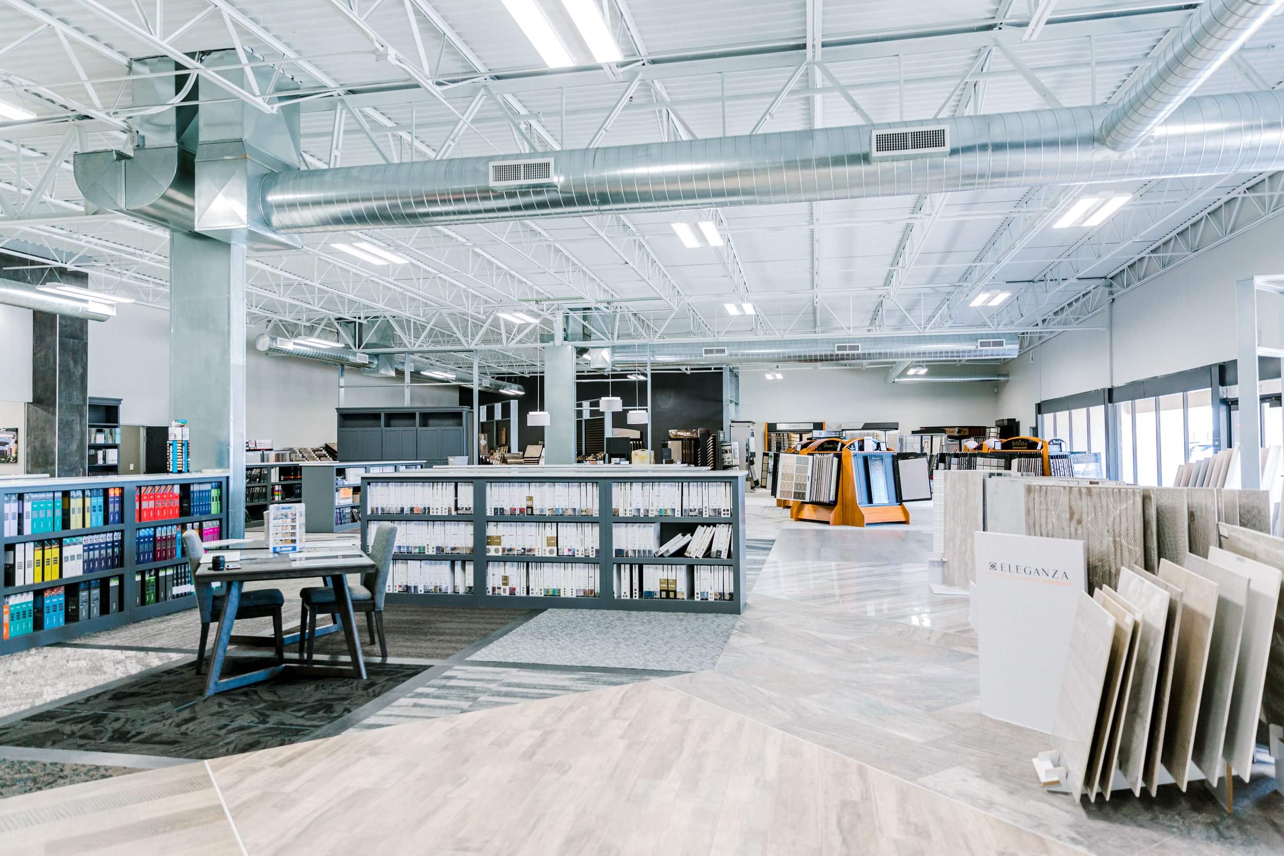 Bright flooring and tile showroom with product displays, sample books, shelves, tables, and exposed industrial HVAC ducts.