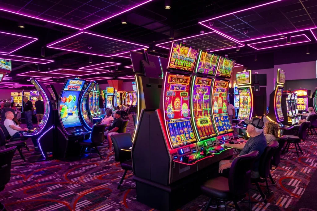 Busy casino floor with colorful slot machines, seated players, and purple neon ceiling lights