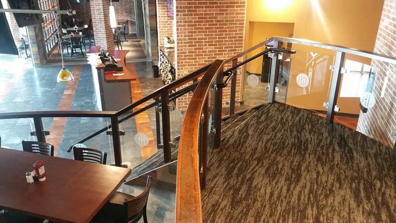 Interior of a modern brick-walled restaurant with a carpeted staircase, glass-and-wood railings, and dining tables.