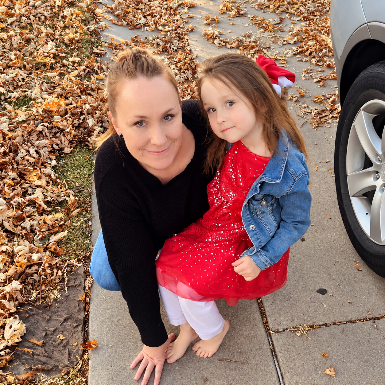Mother and young daughter posing on a leaf-covered sidewalk beside a car, wearing a red dress and denim jacket.