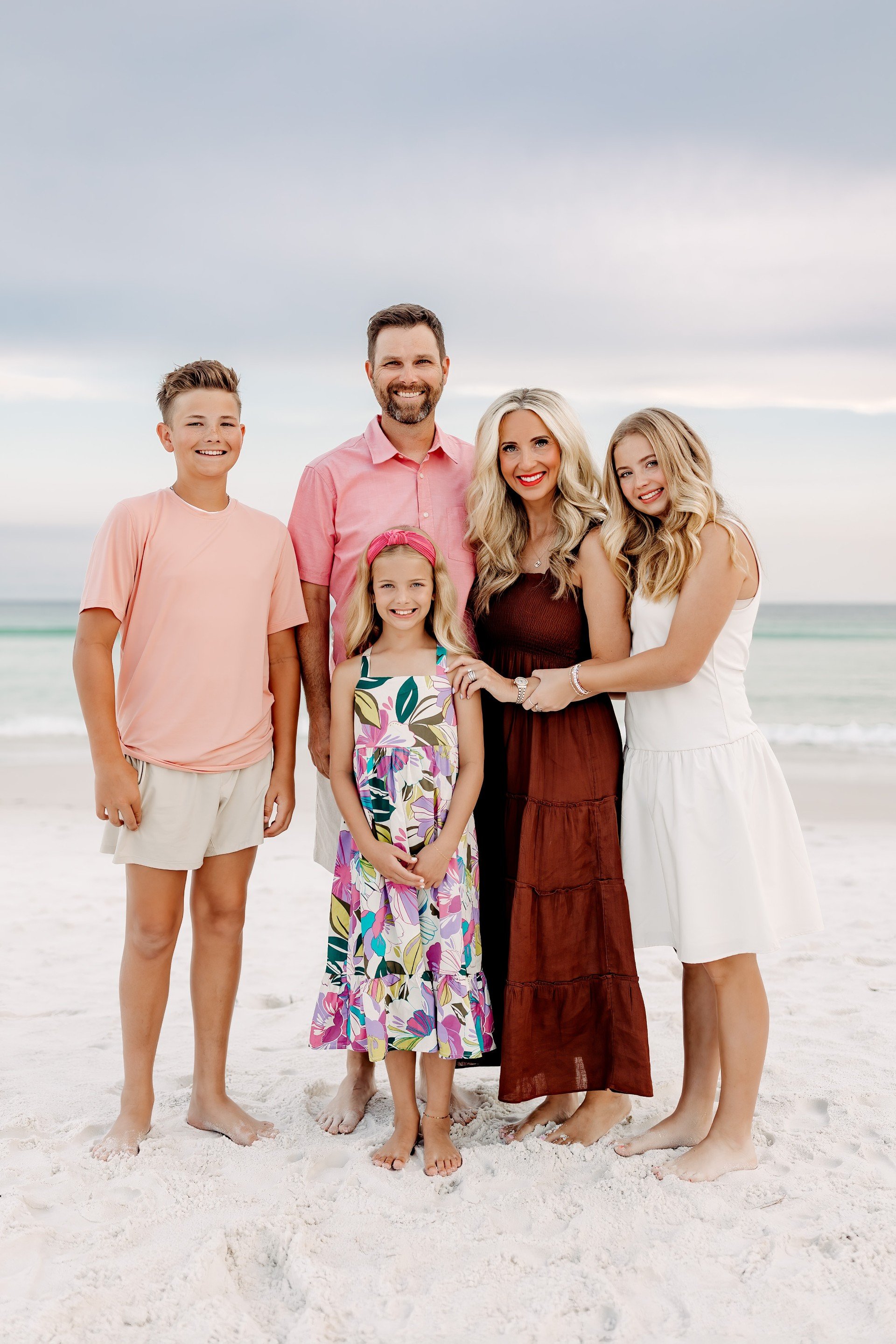 Smiling family of five posing together on a sandy beach beside the ocean