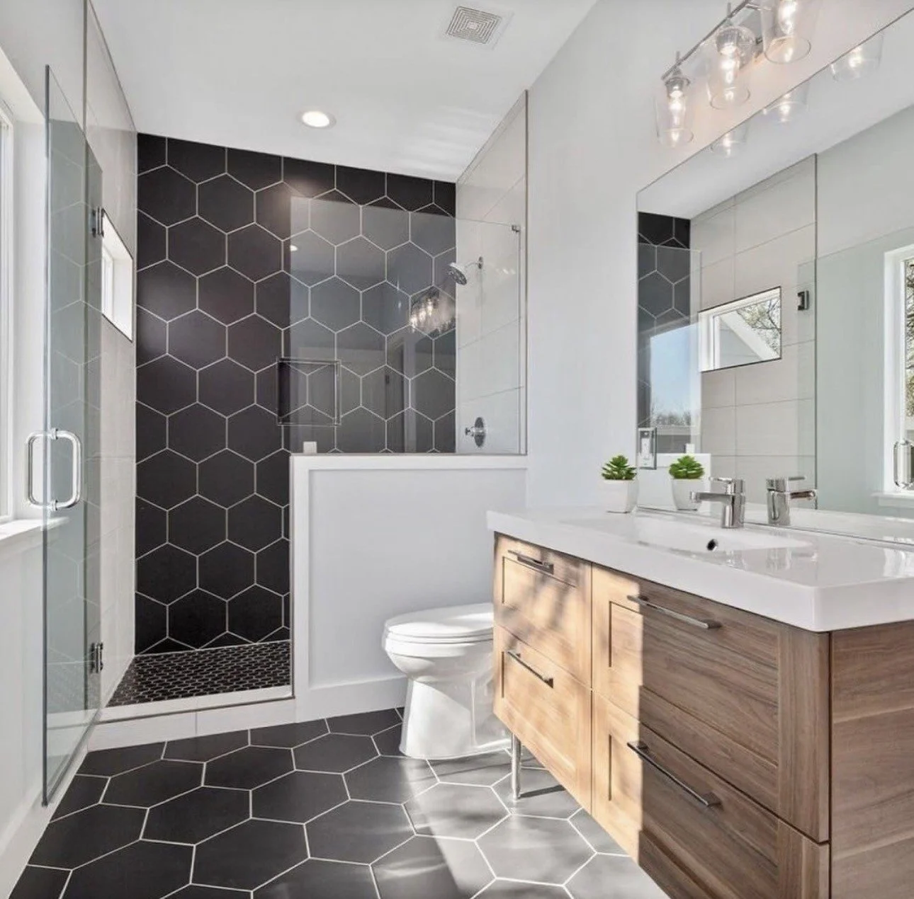 Modern bathroom with black hexagonal tile shower and floor, glass enclosure, white toilet, large mirror, and wood vanity.
