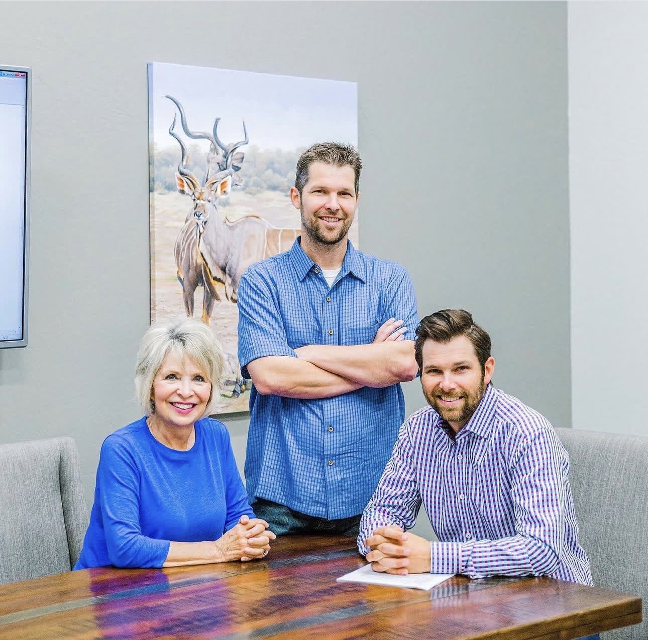 Three smiling coworkers pose around a conference table in an office, with wildlife artwork behind them.