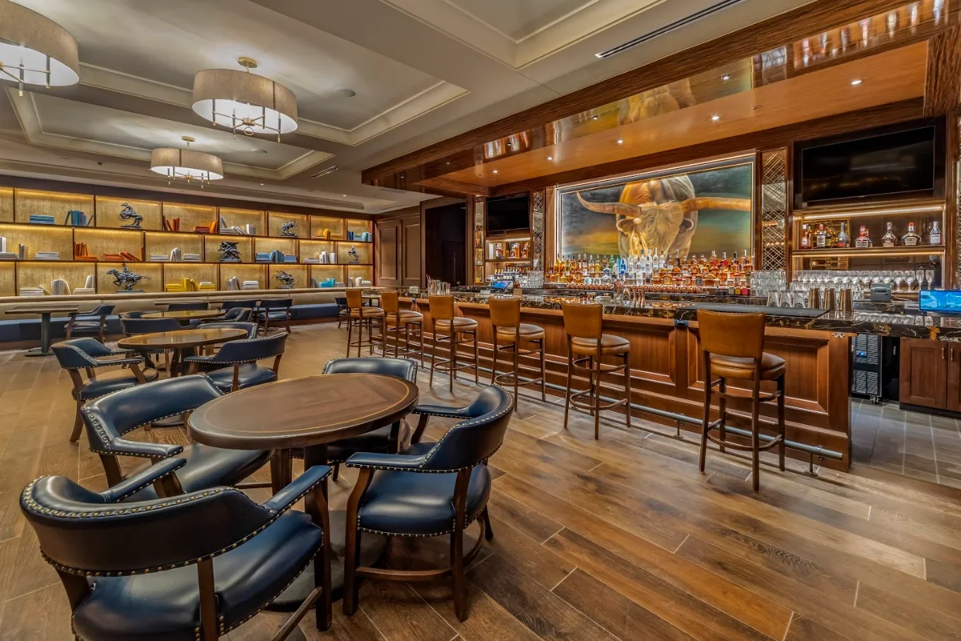 Upscale hotel bar and lounge with leather chairs, wood tables, illuminated bookshelves, extensive liquor display, and longhorn painting