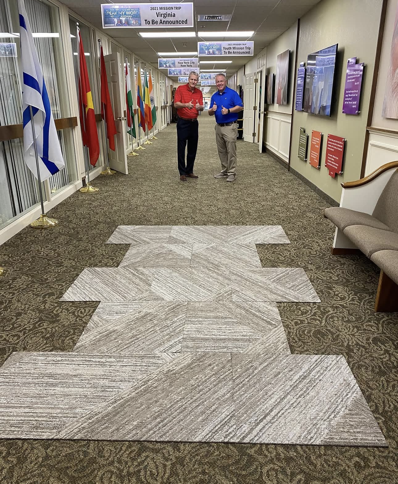 Two men giving thumbs-up in a church hallway lined with international flags, mission trip signs, and geometric gray carpet tiles.