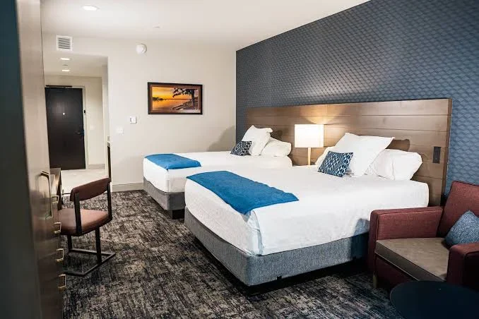Hotel room with two queen beds, blue runners, patterned accent wall, armchair, desk, and nightstands