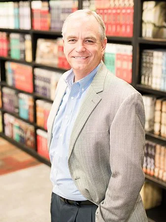 Smiling middle-aged man in a gray blazer standing in a bookstore aisle.