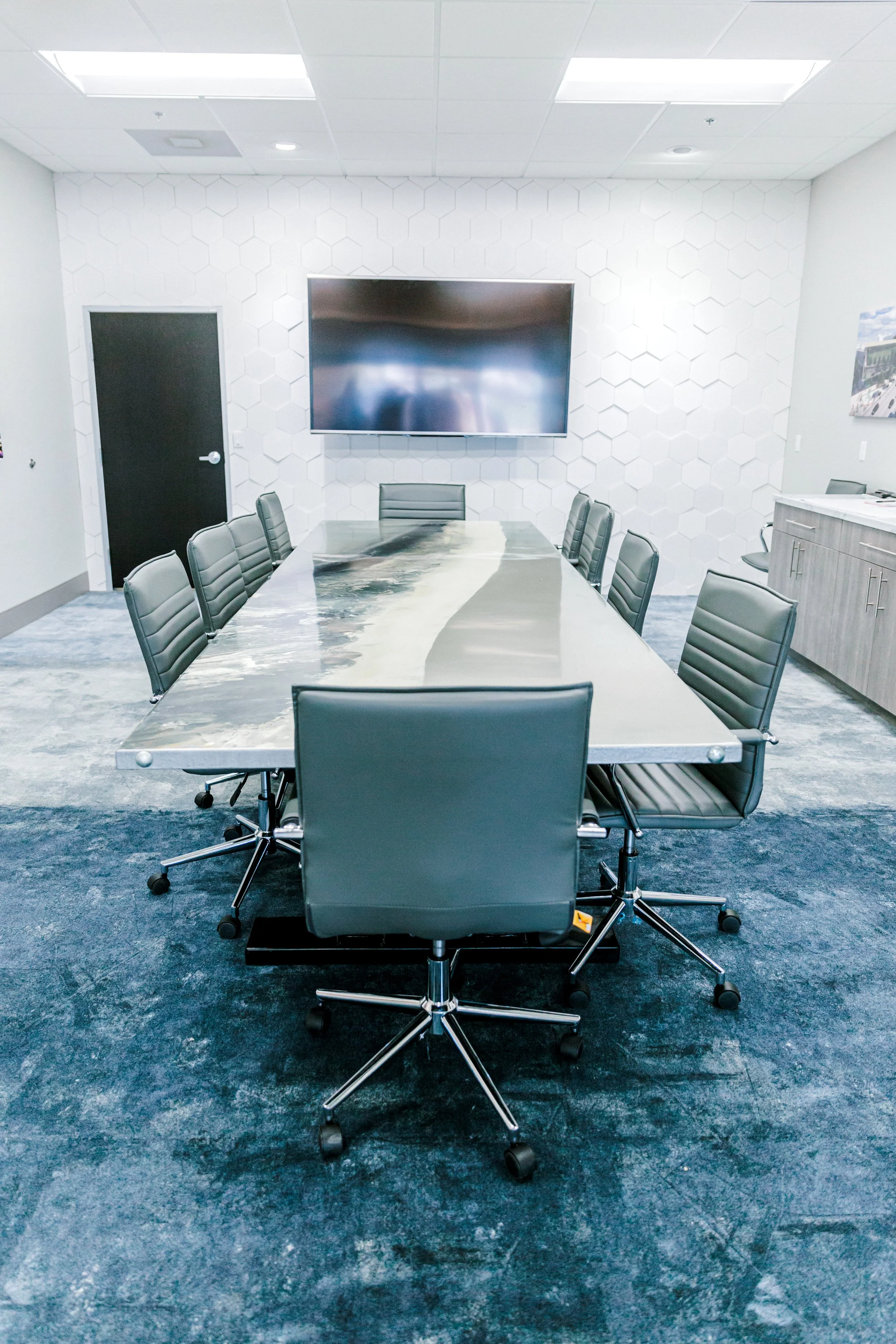 Modern conference room with a long marble table, black leather chairs, wall-mounted TV, and white hexagonal wall paneling