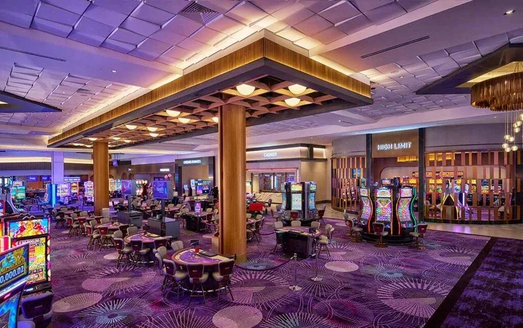Modern casino interior with rows of slot machines, gaming tables, purple patterned carpet, and decorative ceiling lighting