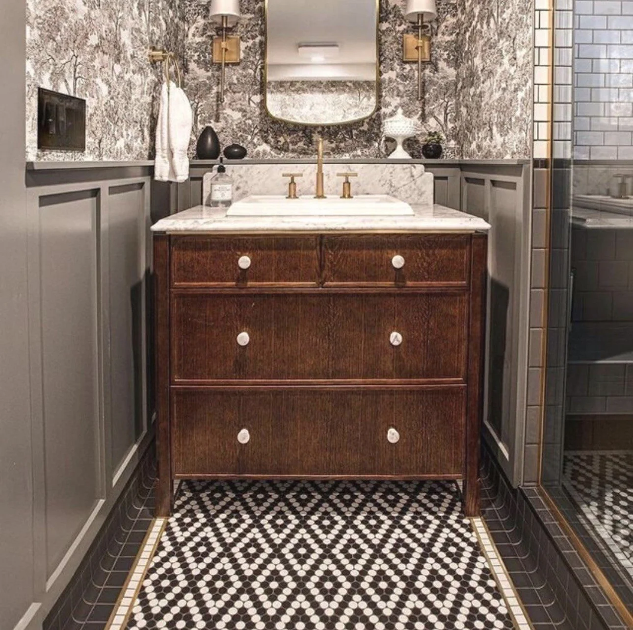 Elegant bathroom vanity with dark wood drawers, marble countertop, brass fixtures, patterned wallpaper, and black-and-white mosaic tile floor