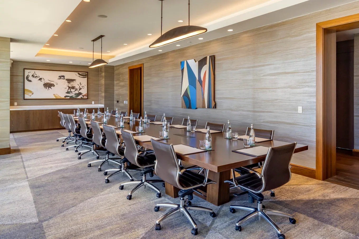Modern corporate conference room with a long boardroom table, leather swivel chairs, bottled water, artwork, and pendant lighting