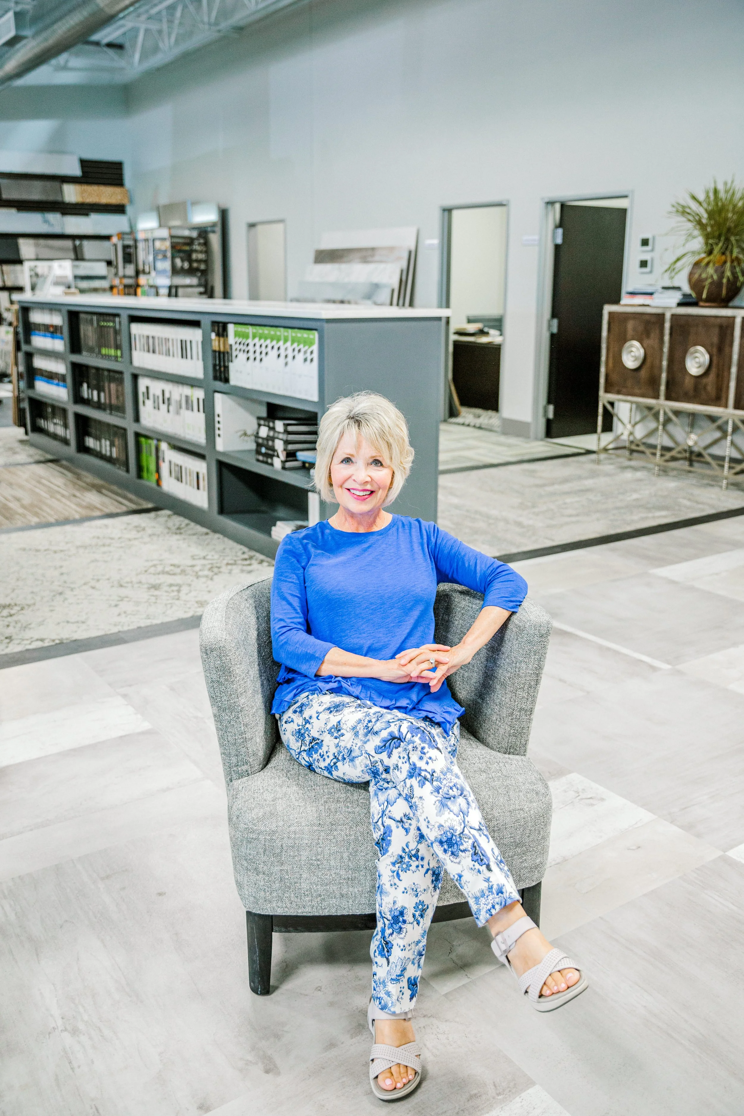 Smiling older woman in a blue top and floral pants sitting in a modern office armchair.