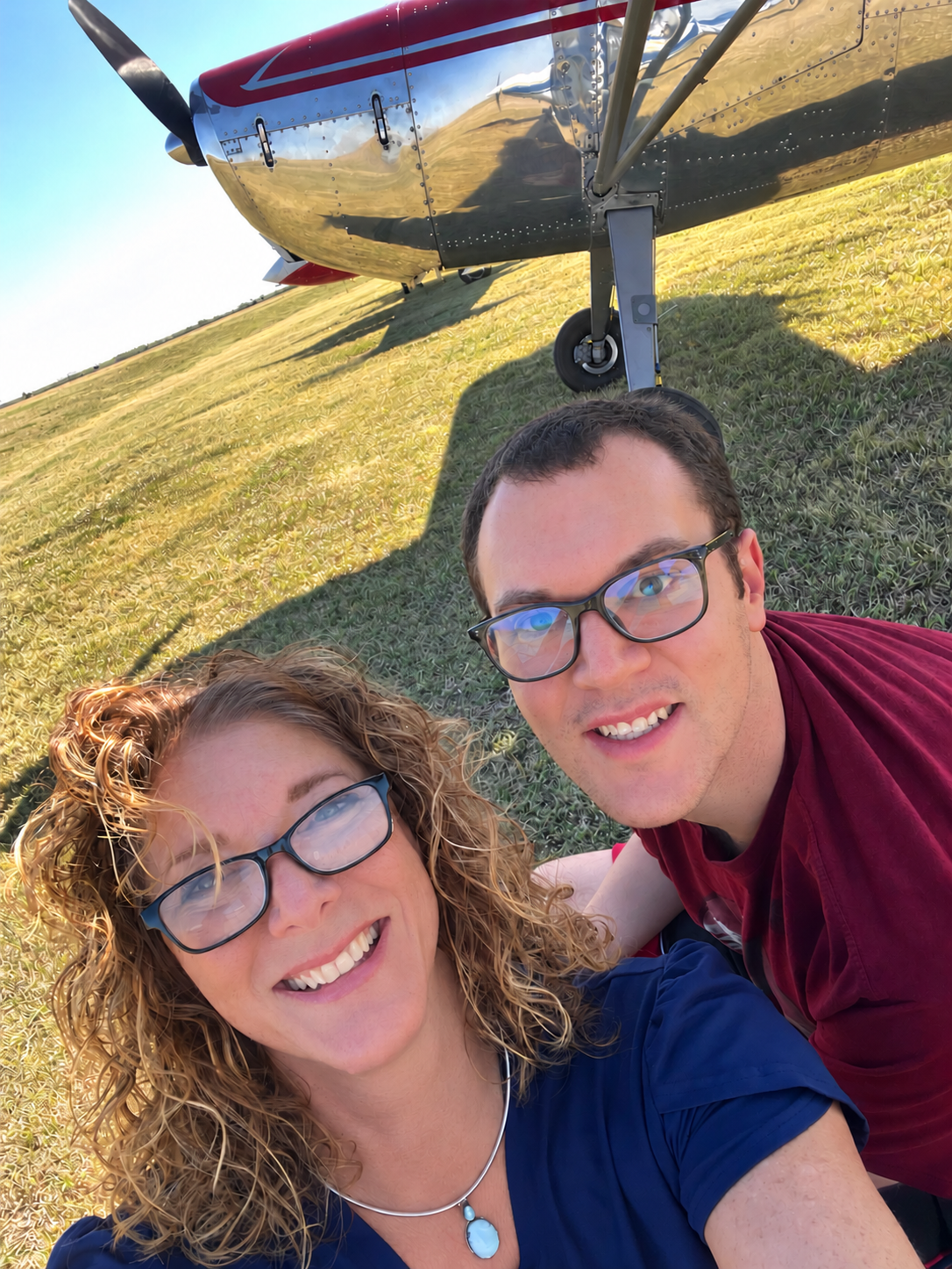 Smiling couple taking a selfie beside a polished vintage airplane on a grassy airfield