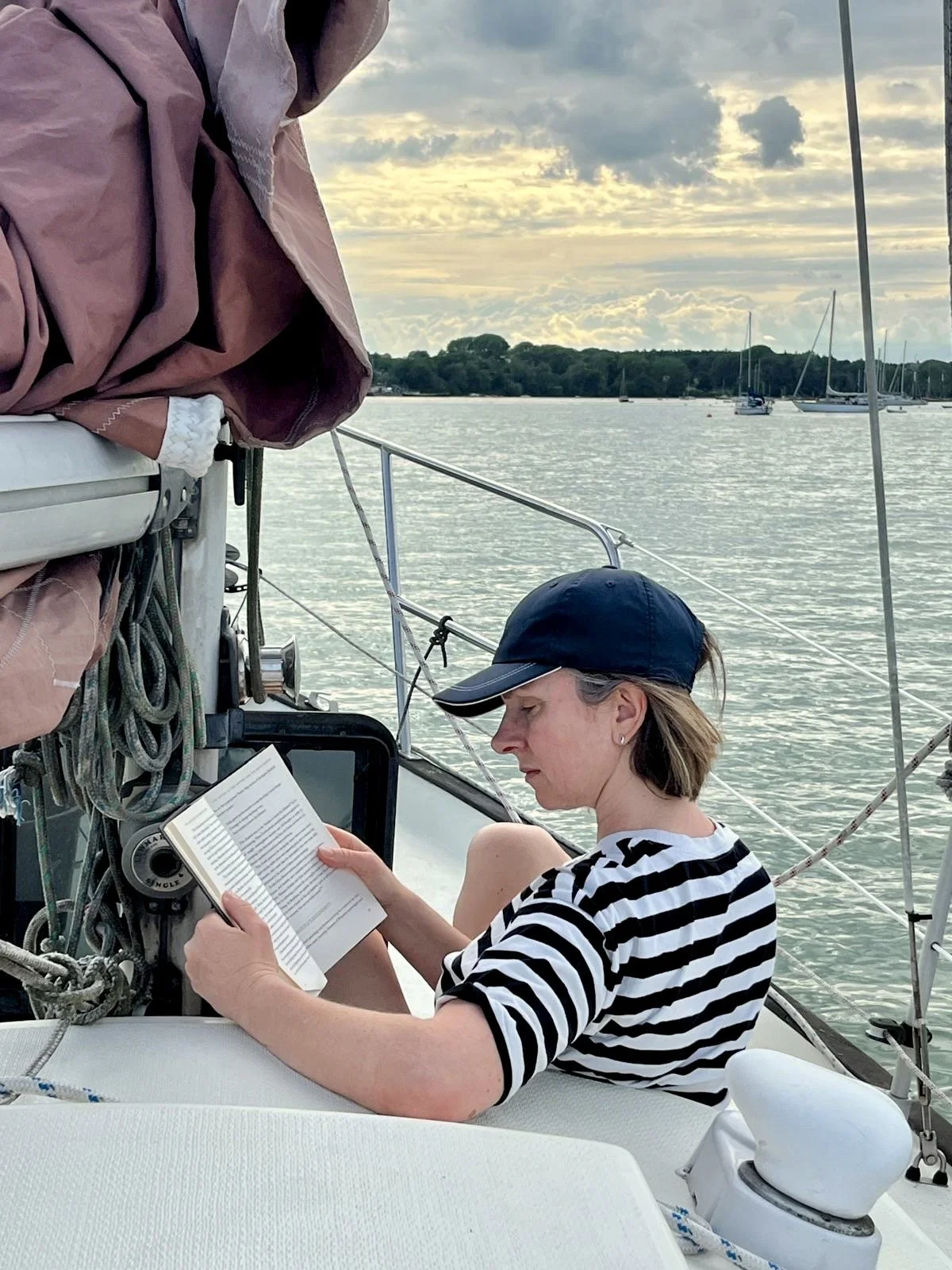 A woman reading a book on a boat, with sailboats and water in the background.