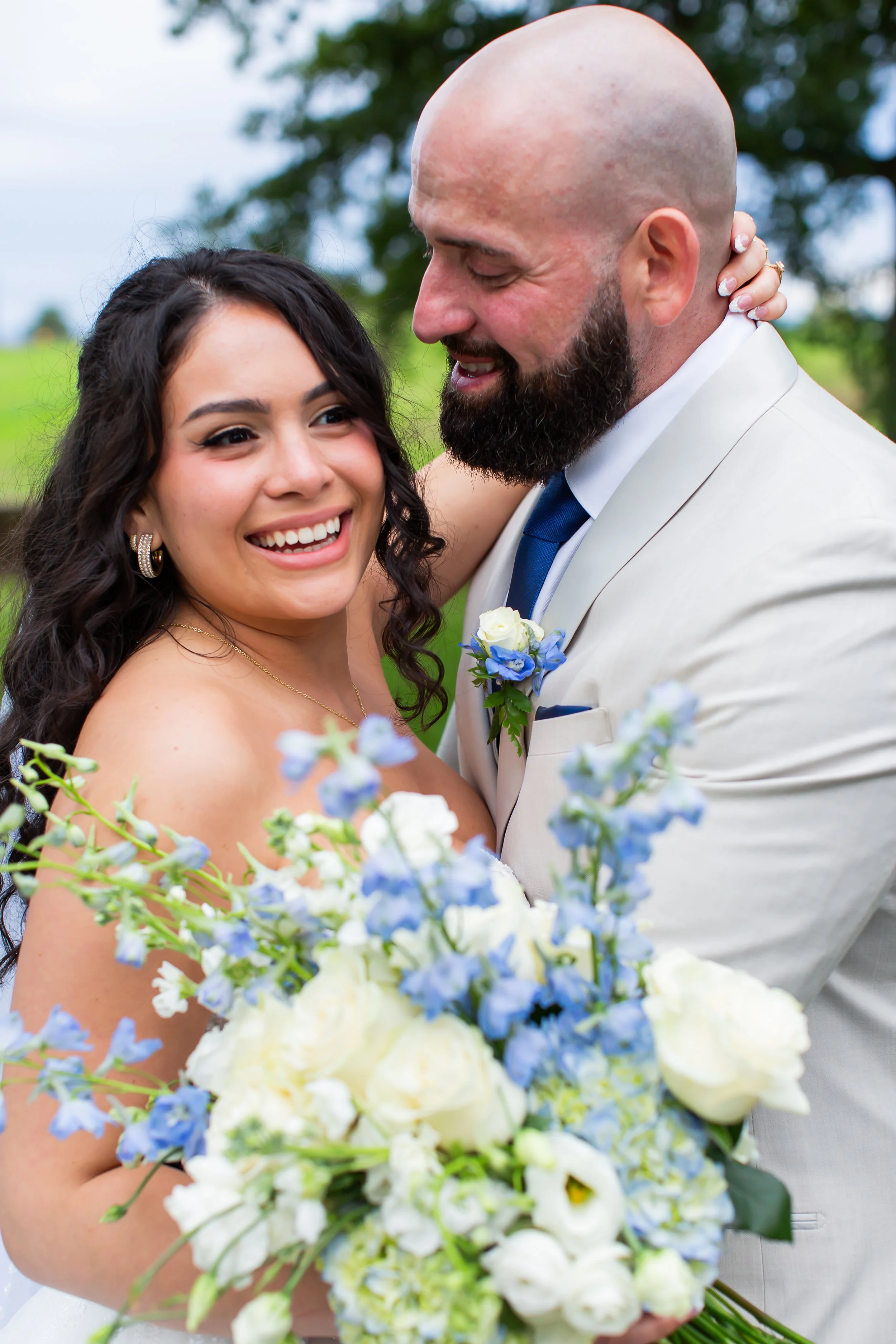 Close-up photo of a newlywed couple embracing outdoors, with bride holding a bouquet of white and blue flowers, both smiling and looking happy, surrounded by greenery.