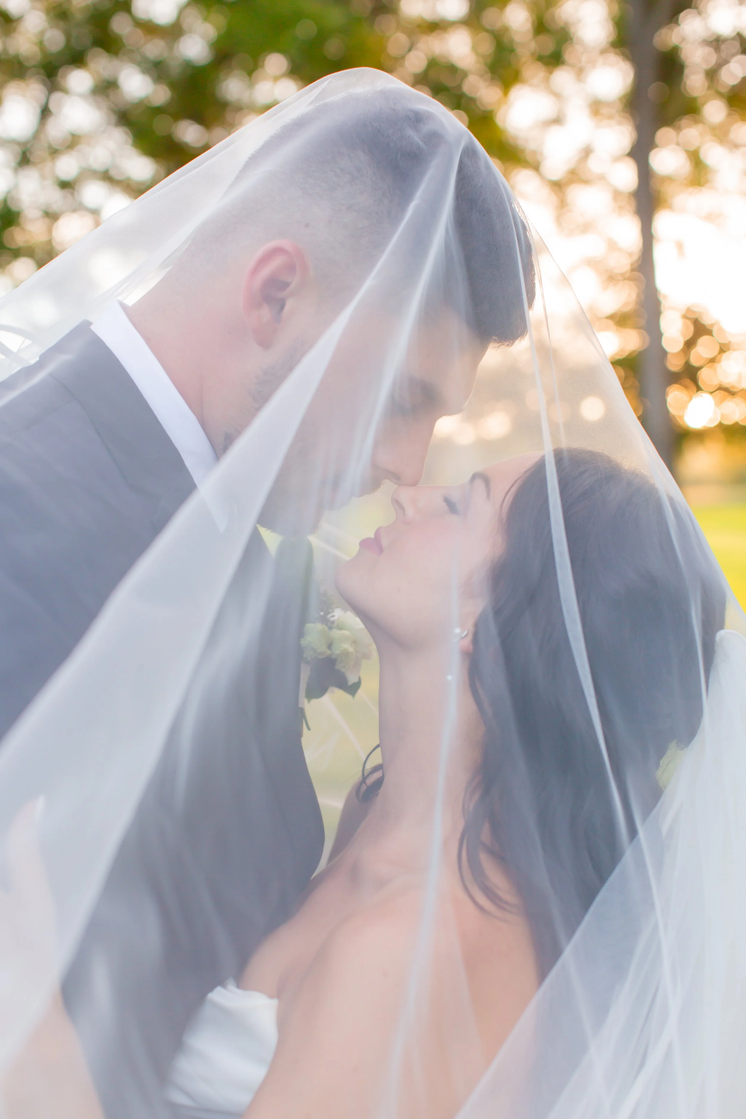 A bride and groom under a wedding veil, leaning close with foreheads touching, during an outdoor wedding. The background shows trees with warm sunlight.
