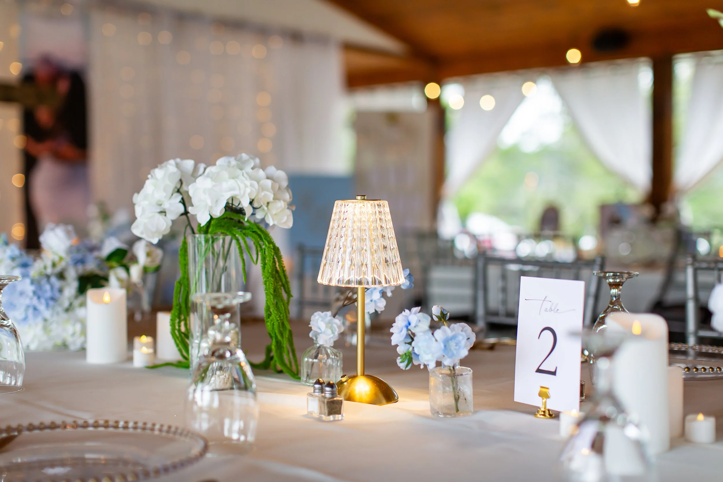 Elegant indoor event space with a table centerpiece of white flowers and a decorative lamp, surrounded by various candles and table cards, with large windows and white curtains in the background.