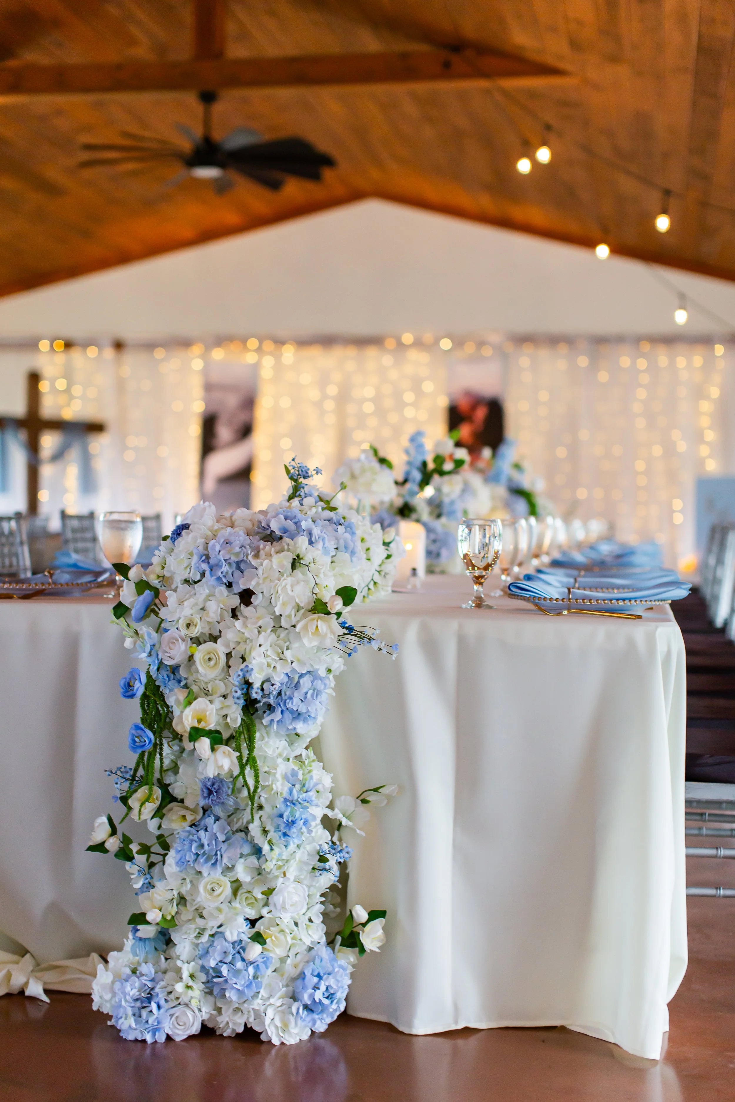 A table decorated with a white tablecloth, floral arrangements in shades of white and light blue, with glassware and place settings, set for a wedding or celebration. In the background, there are string lights and a cross, suggesting an indoor or semi-outdoor venue with a religious or formal theme.
