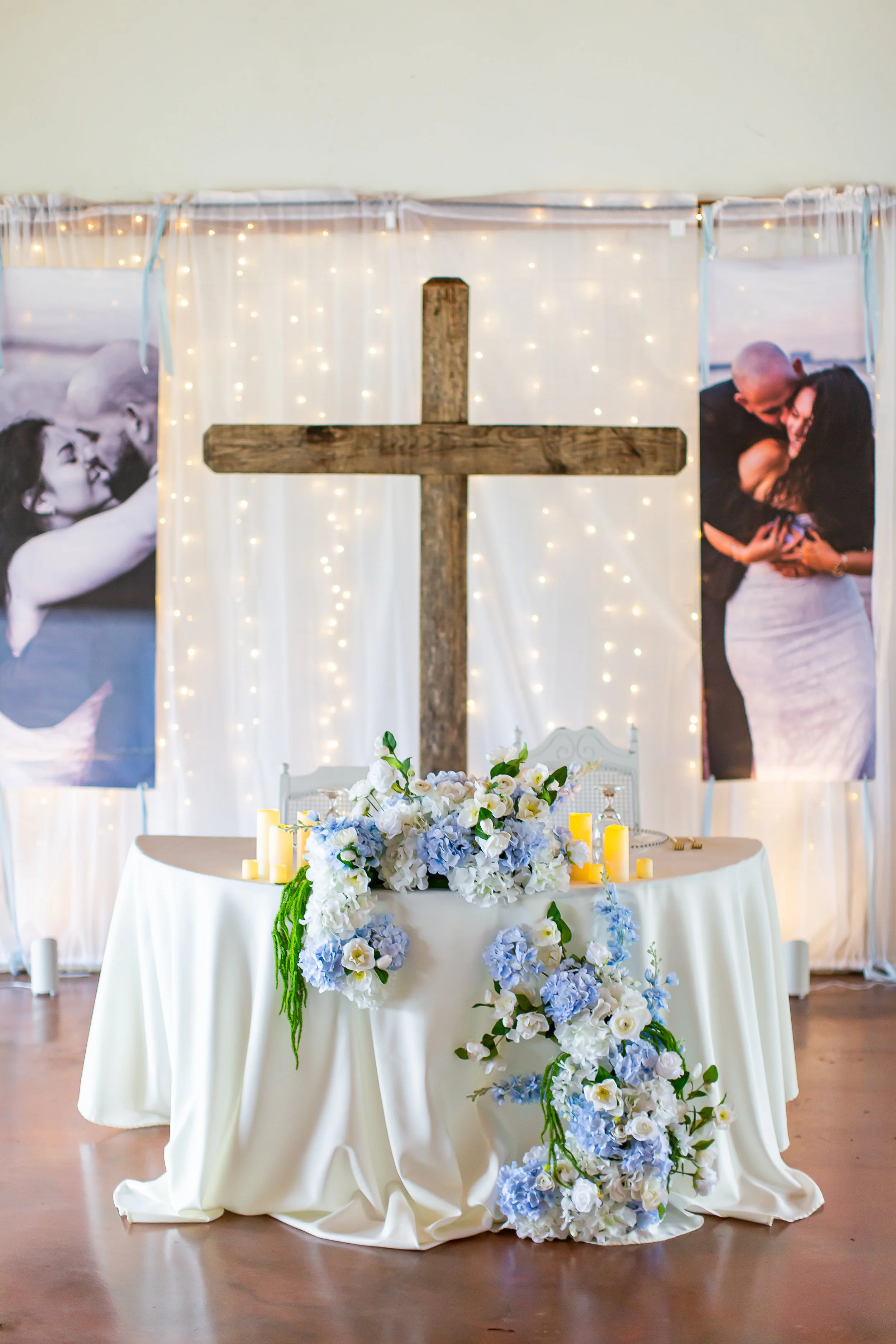A table covered with a white cloth, decorated with white and blue flowers and candles, set in front of a backdrop with a cross, fairy lights, and large black and white photographs of a couple embracing.