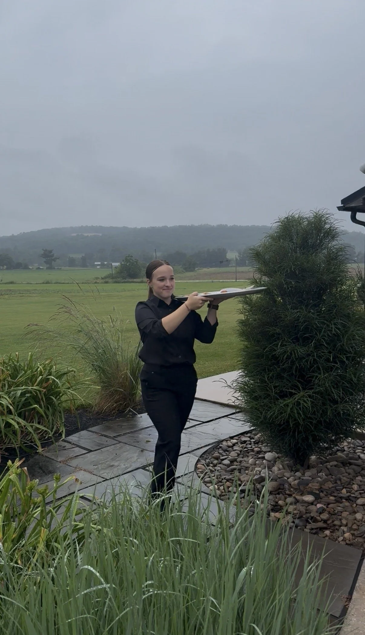 A woman dressed in black holding a tray with food outdoors on a cloudy day, with green fields and hills in the background.