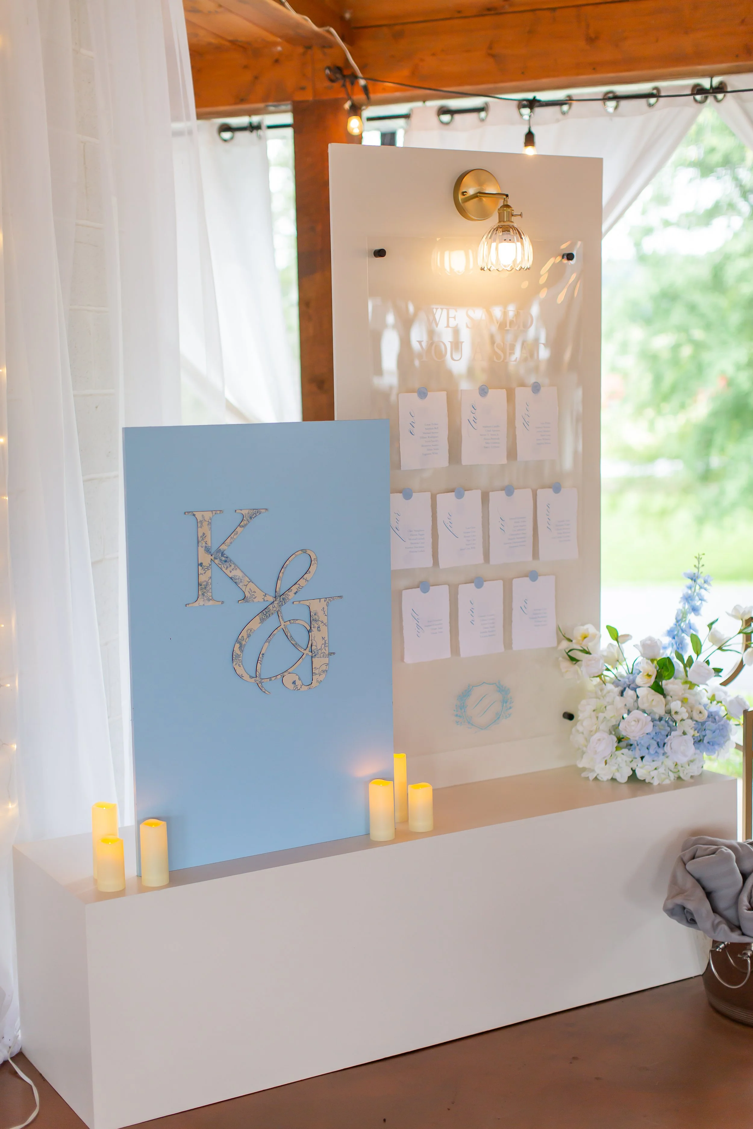 A wedding or event seating chart display with a blue sign featuring the initials 'K & J' and a white board with handwritten seating arrangements, decorated with flowers and candles.