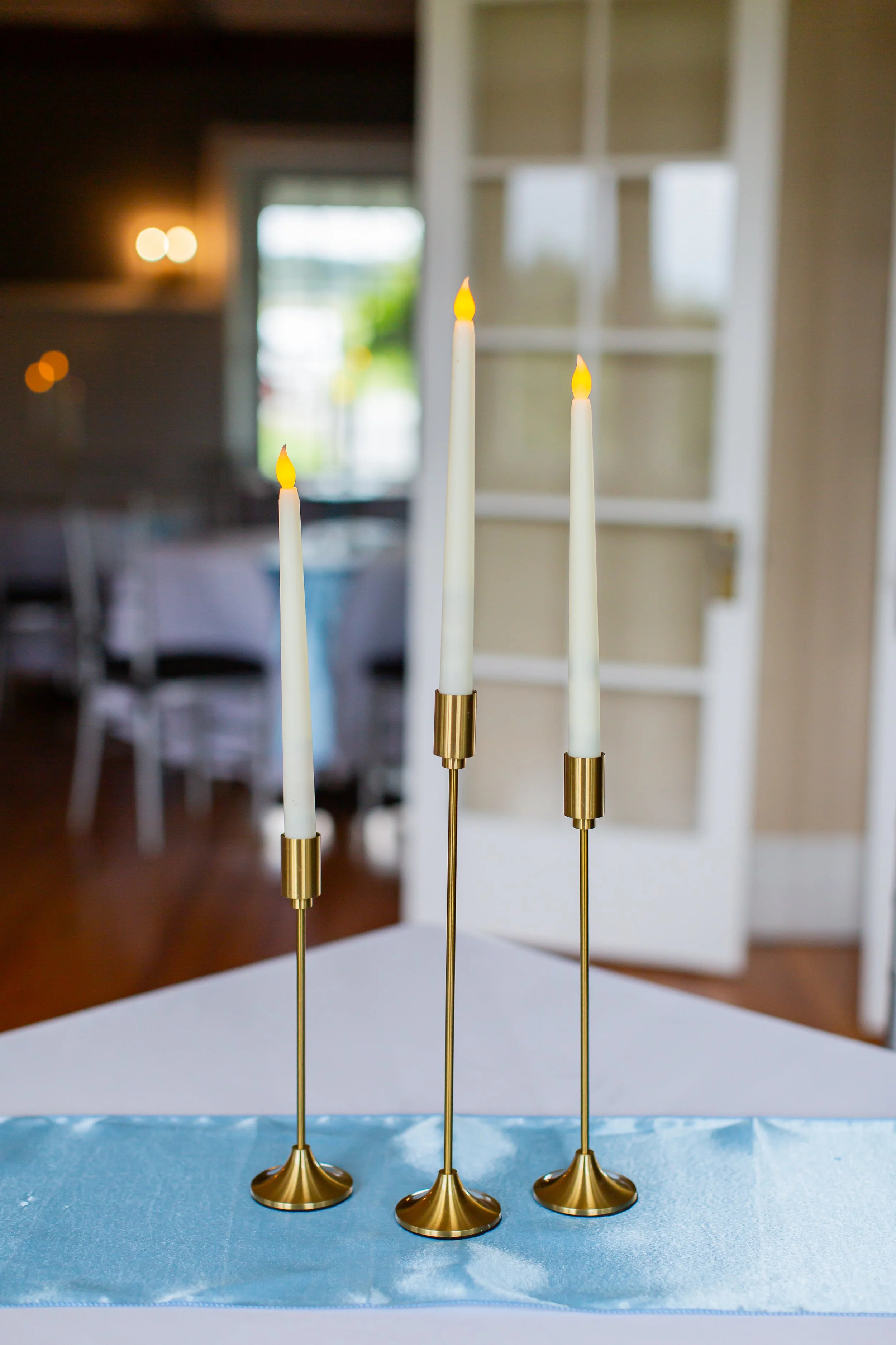 Three tall, slim, gold candleholders with white candles topped with small flames, placed on a table with a blue table runner in a room with windows and shelves in the background.