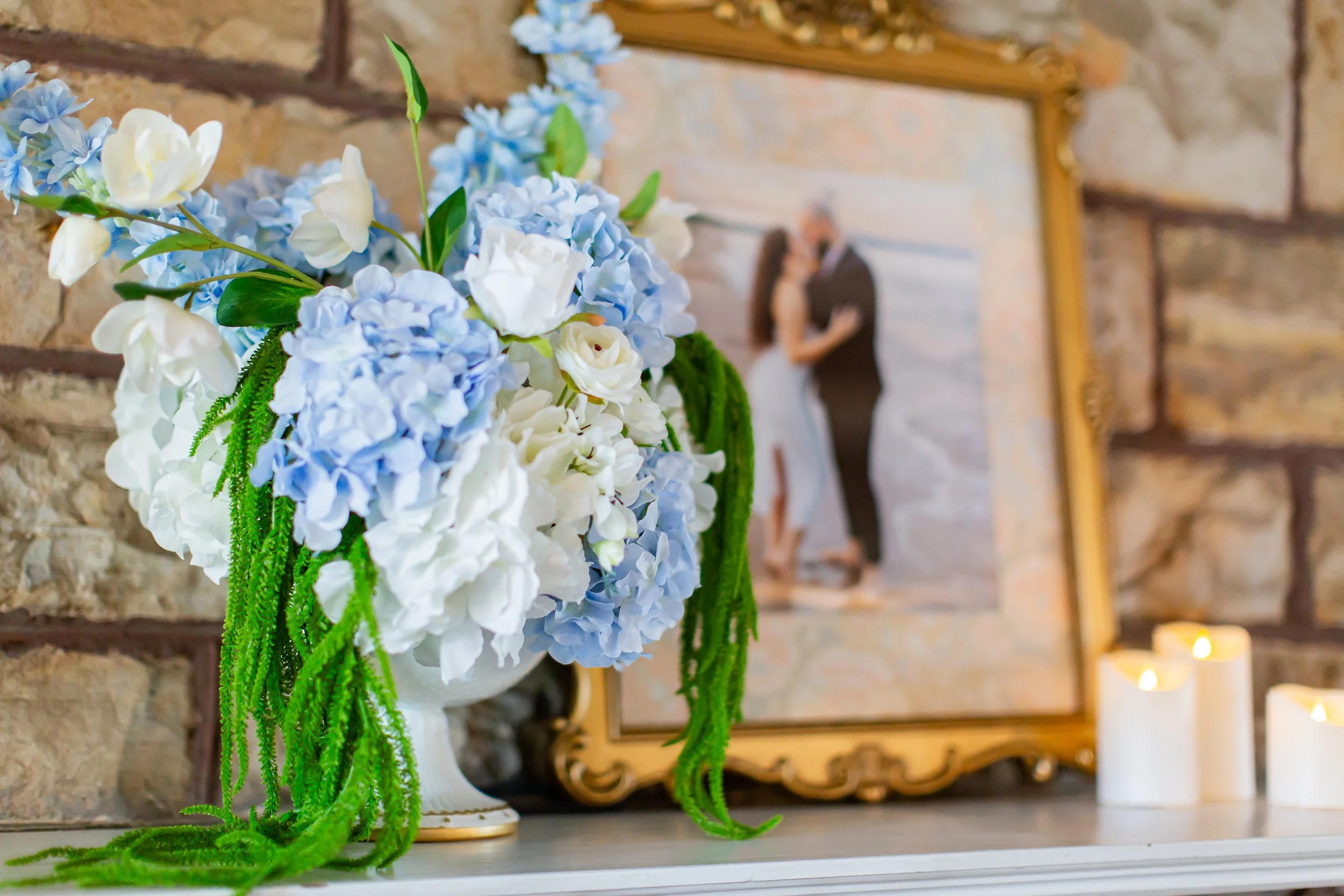 A floral arrangement featuring white and light blue hydrangeas with green foliage in a white vase, placed on a white surface with a blurred background of a brick wall, gold framed mirror, and candle.