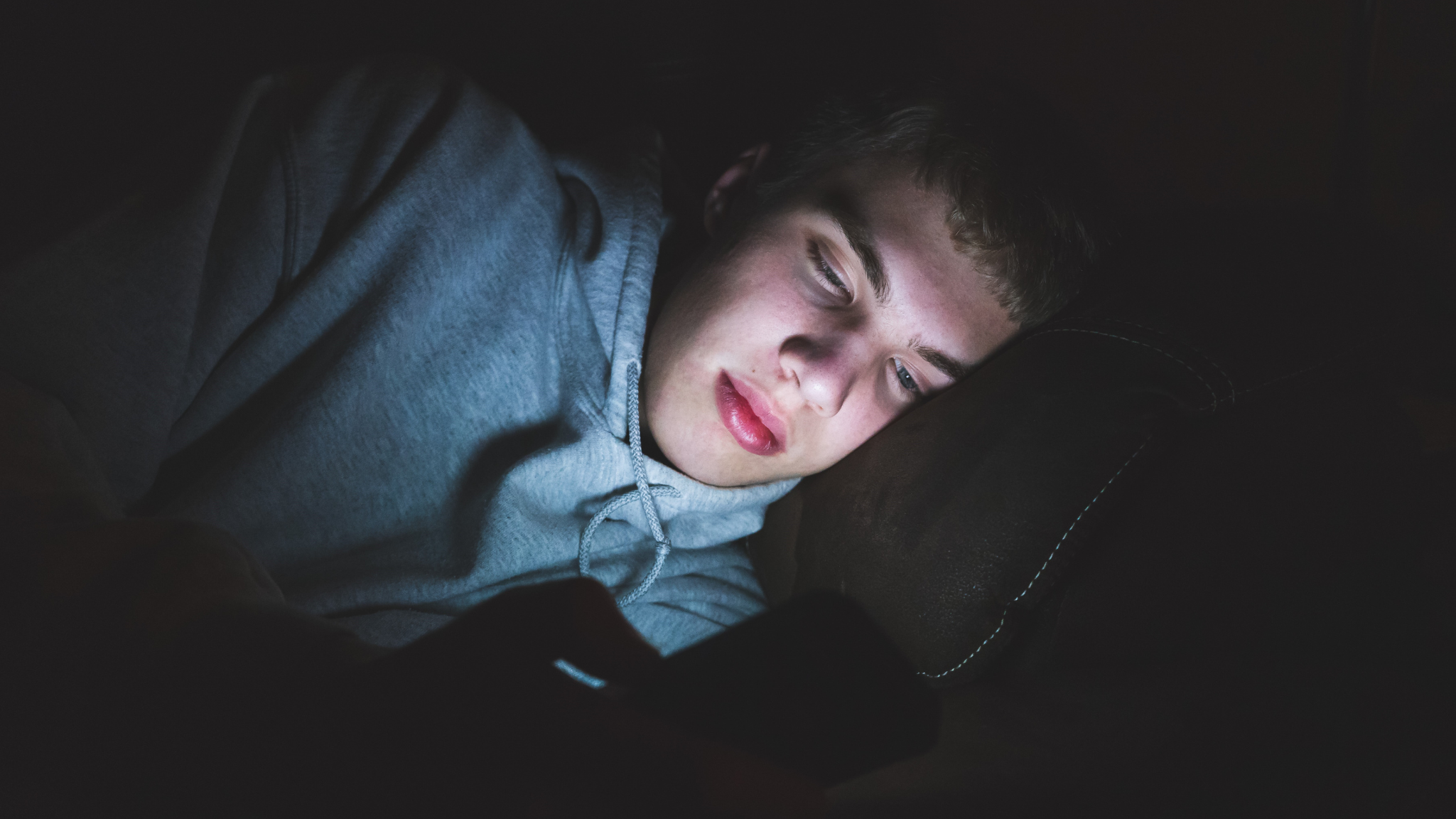 Young man lying on a dark couch, resting his head on a black cushion, wearing a gray hoodie, looking at a screen with a focused or concerned expression.