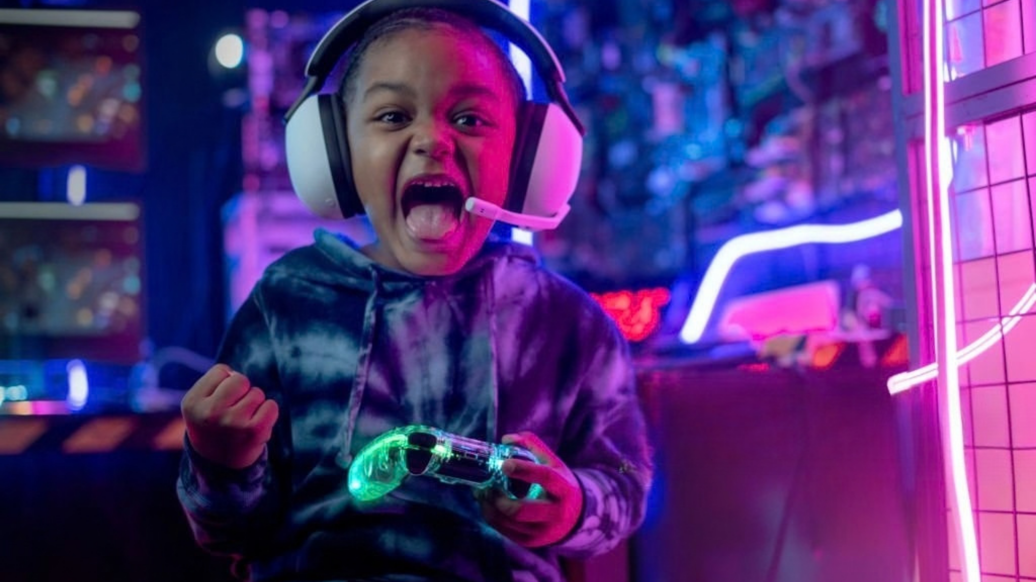 A young boy with a headset and glowing controller excitedly making a fist in a neon-lit arcade or gaming center.