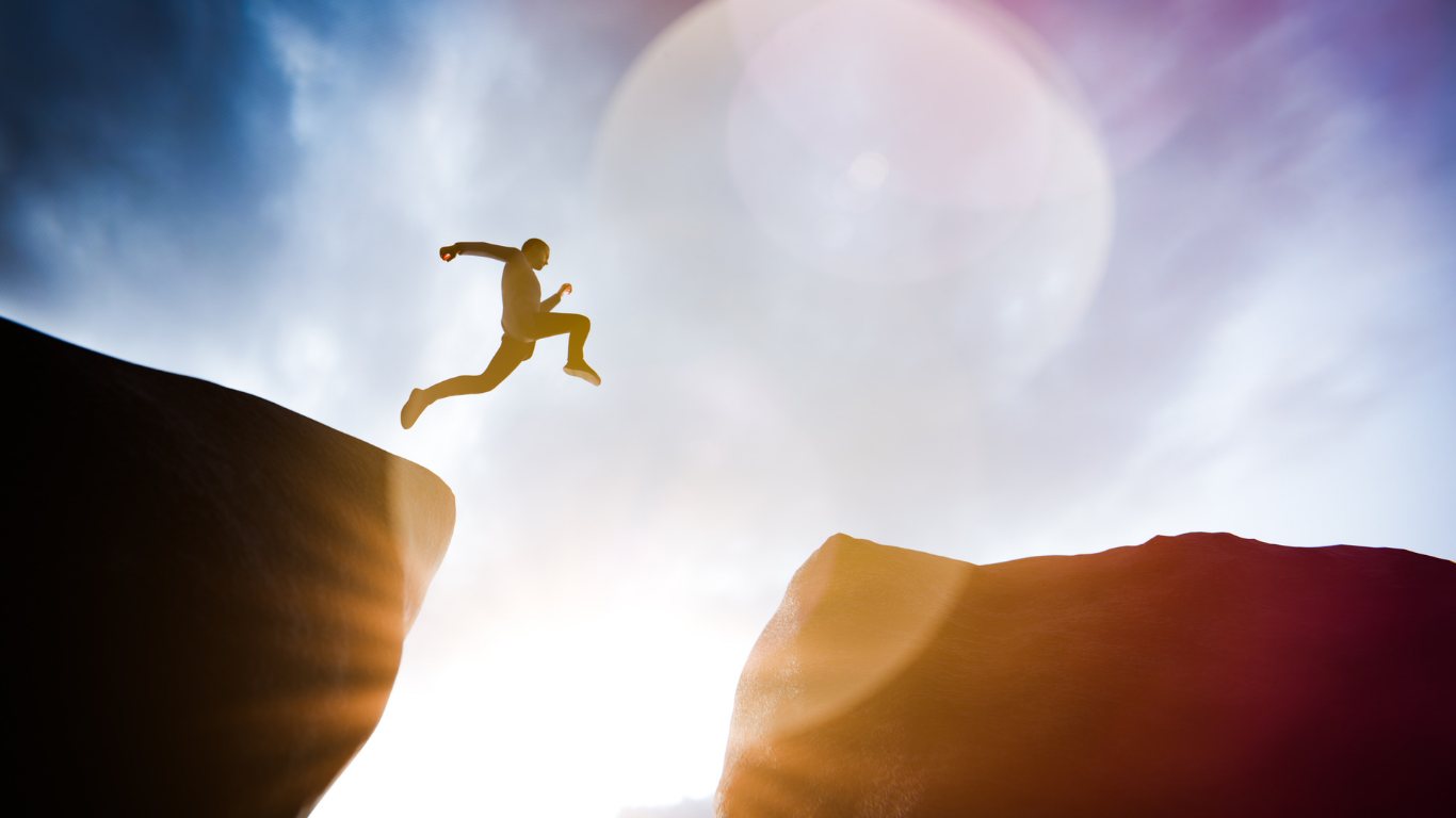 Silhouette of a man in a suit leaping across a gap between two rocks against a bright sky with lens flare.