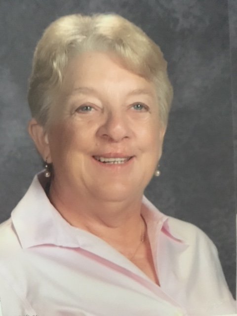 A portrait of an older woman with short blonde hair, smiling, wearing a light pink collared shirt and pearl earrings.