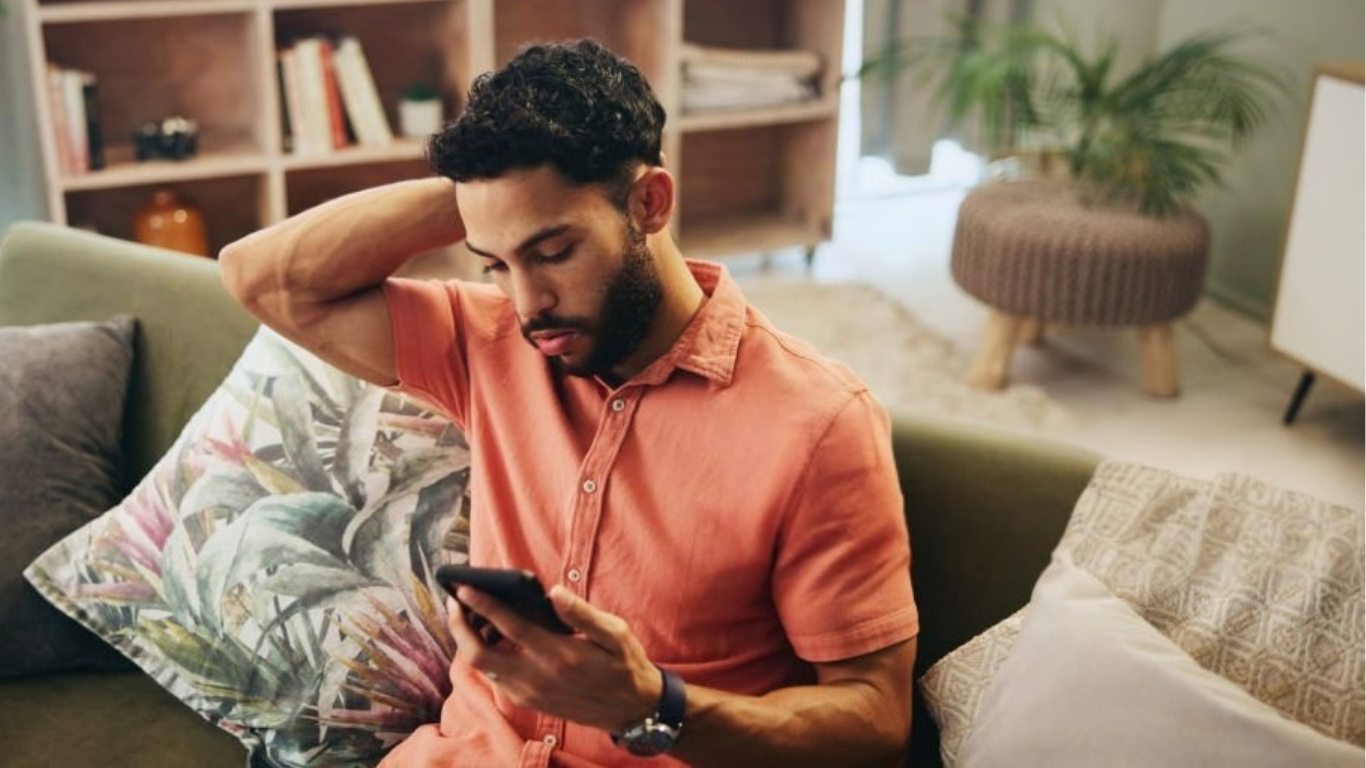 A young man with dark, curly hair and a beard sits on a sofa, looking down at his smartphone with a worried expression. He is wearing an orange collared shirt and a wristwatch. The background includes a bookshelf with books, a potted plant, and a cozy living room setting.
