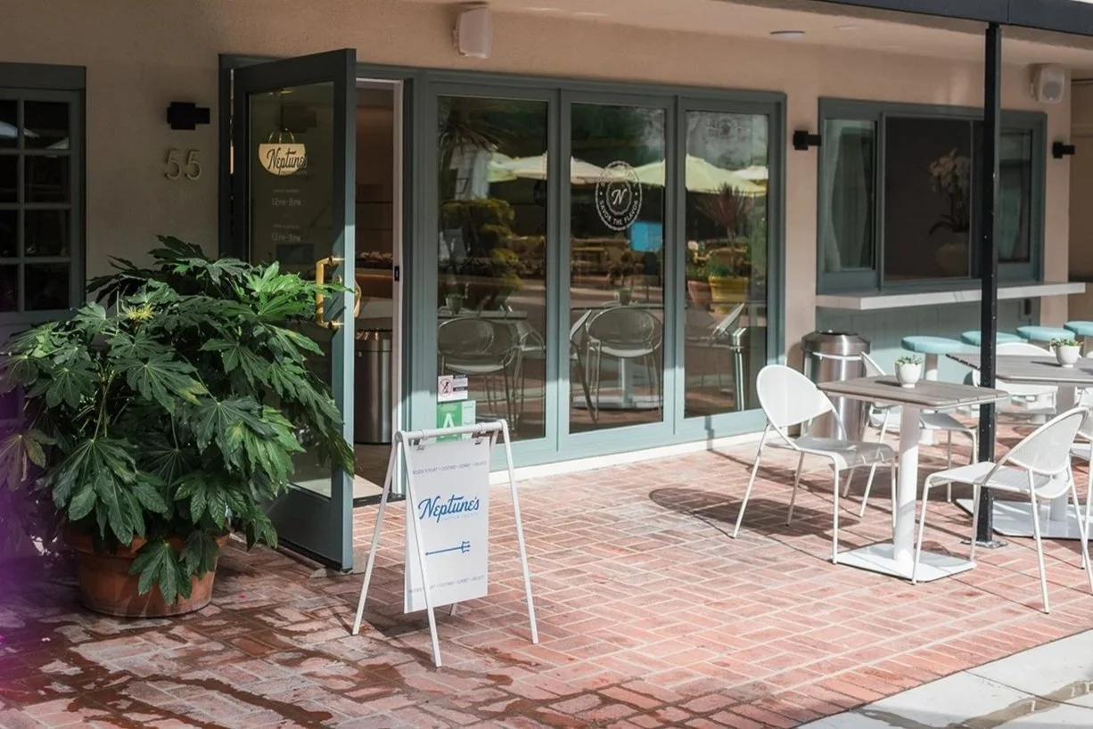 Outdoor seating area of a café with white tables and chairs, potted plants, and a glass door entrance with a sign reading 'Neptunes'