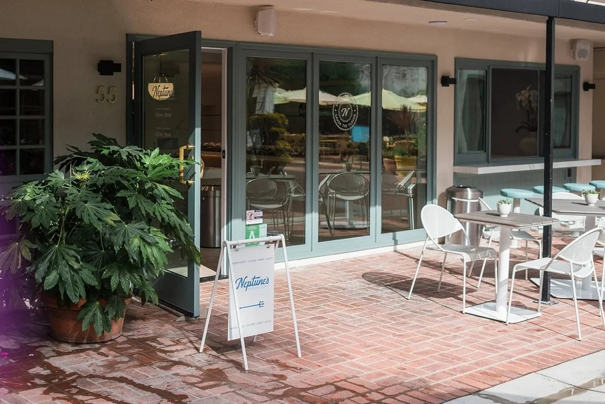 Outside patio area of a restaurant with white tables and chairs, potted plants, and a large leafy plant in a pot. The door has a sign for Neptune's, and the restaurant. Reflective glass doors show the outdoor scene.
