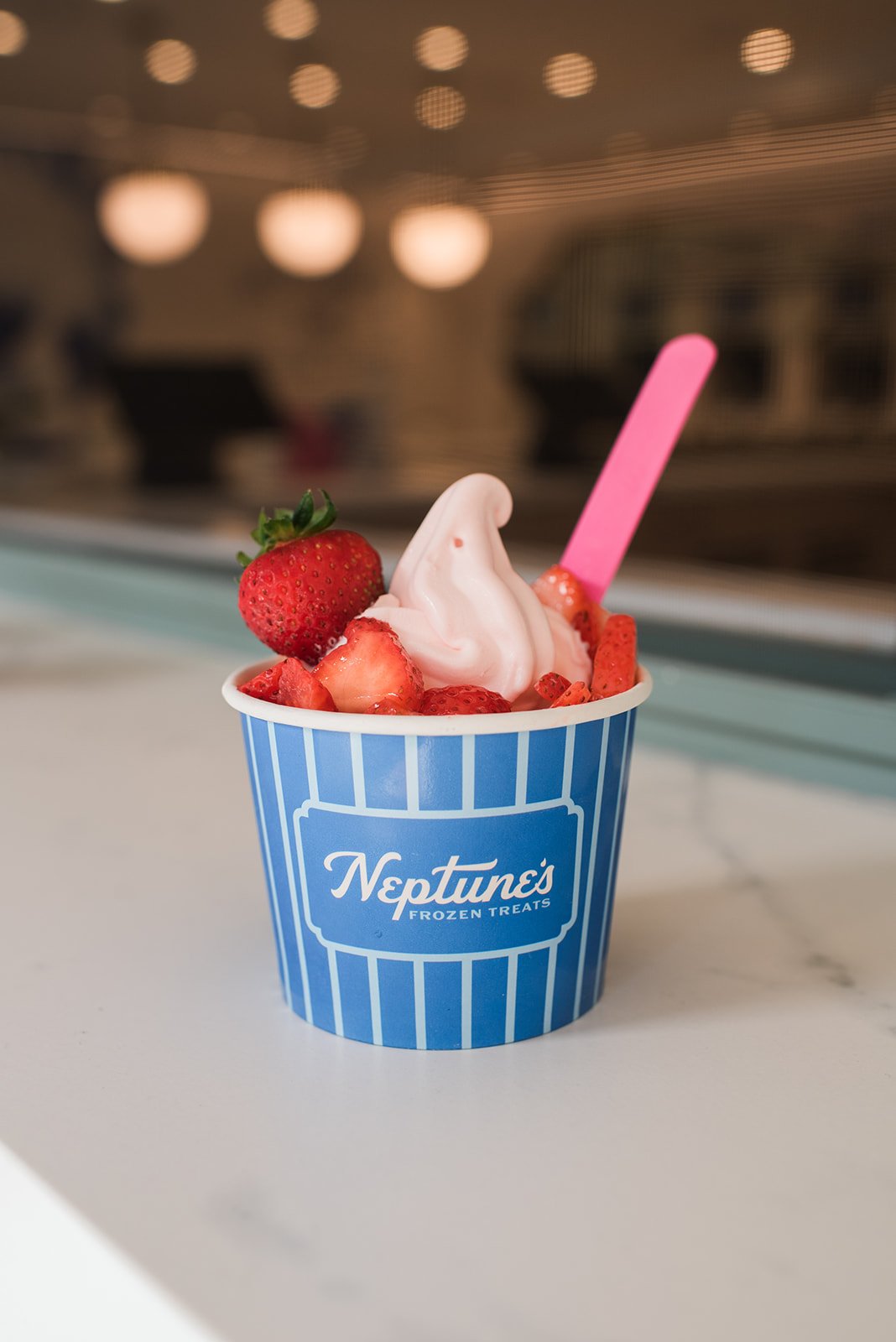 Frozen yogurt cup with pink soft serve, topped with sliced strawberries and a whole strawberry, pink spoon, in a blue striped cup labeled 'Neptune's Frozen Treats', on a white surface with blurred background.
