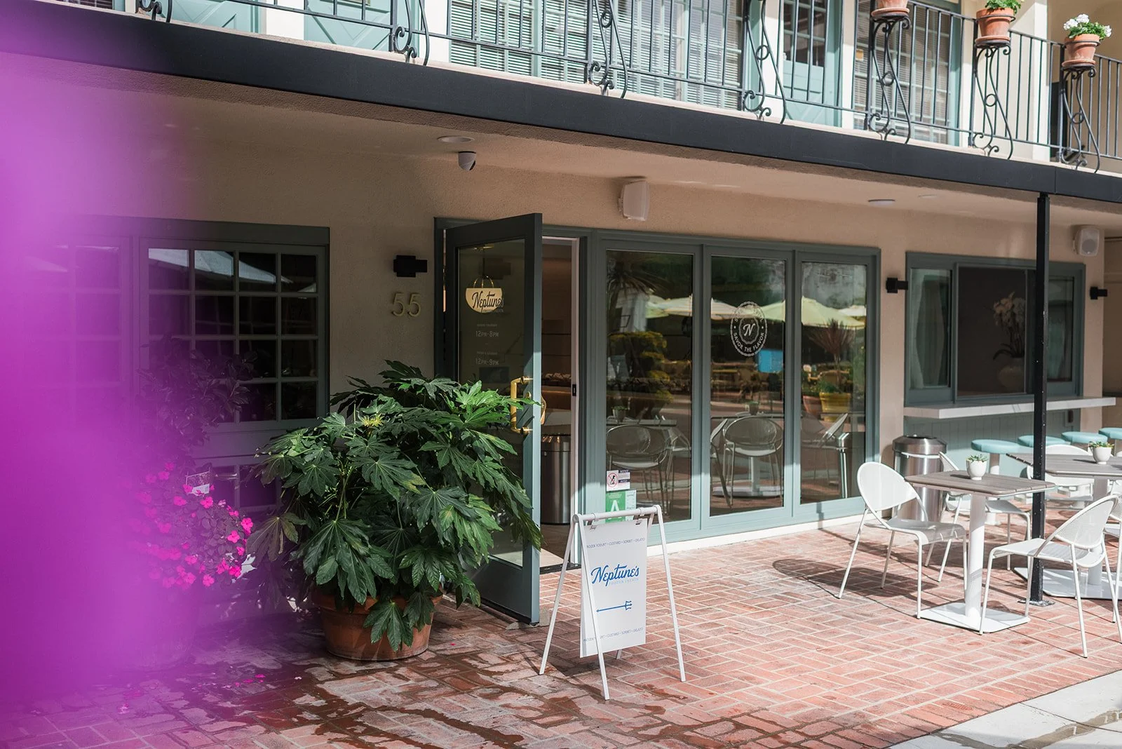 Exterior view of a restaurant named Neptune's with outdoor seating and large potted plants; a white metallic table and chairs are on a brick patio, with a glass door entrance and window, and a sign indicating business hours and location.