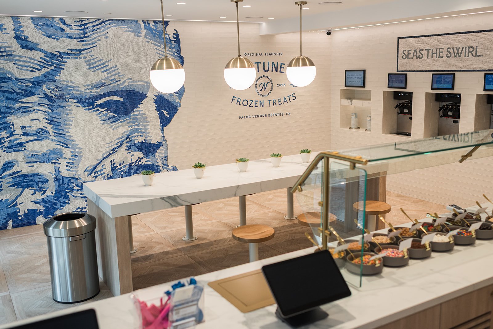 Frozen yogurt shop with a mosaic wall of a woman's face, various frozen treats, small potted succulents, and seating area with three pendant lights.