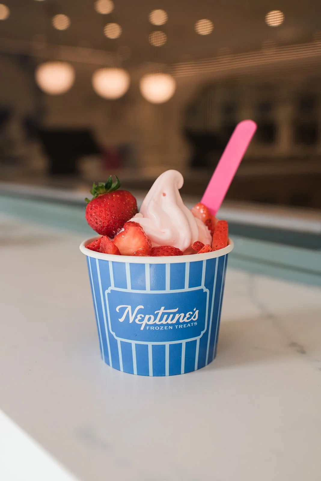A cup of frozen yogurt topped with strawberries and a pink spoon, in a blue and white striped cup from Neptune's Frozen Treats.