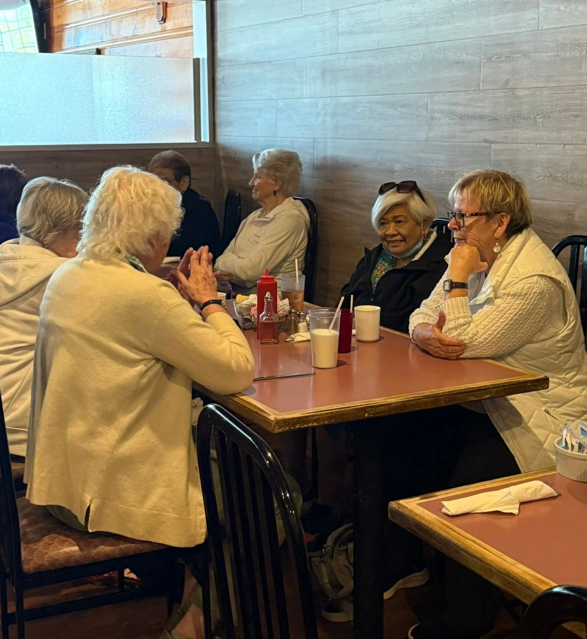 A group of elderly women sitting around a table in a restaurant, engaging in conversation, with drinks, condiments, and napkins on the table.