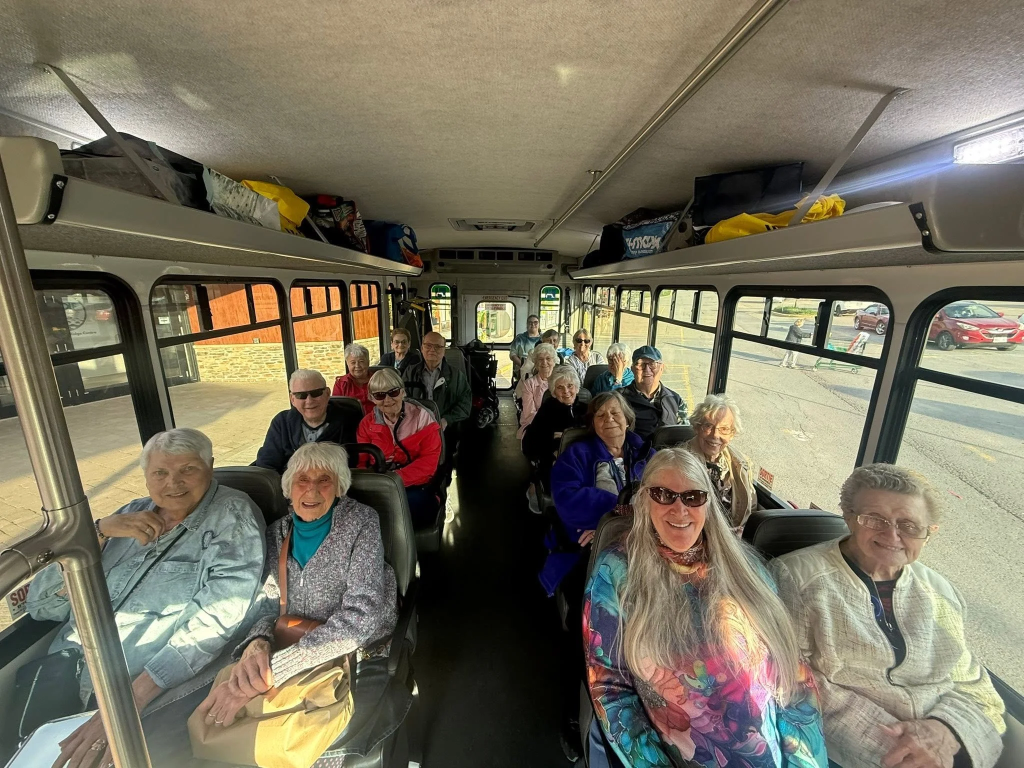 Group of elderly people sitting on a bus, smiling and enjoying a group outing, outside view shows parked cars.