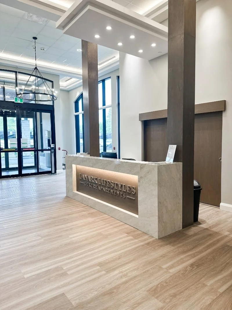 Reception area with a marble countertop sign that reads "Lanark Lifestyle's Luxury Apartments," large windows, and a glass entrance door.