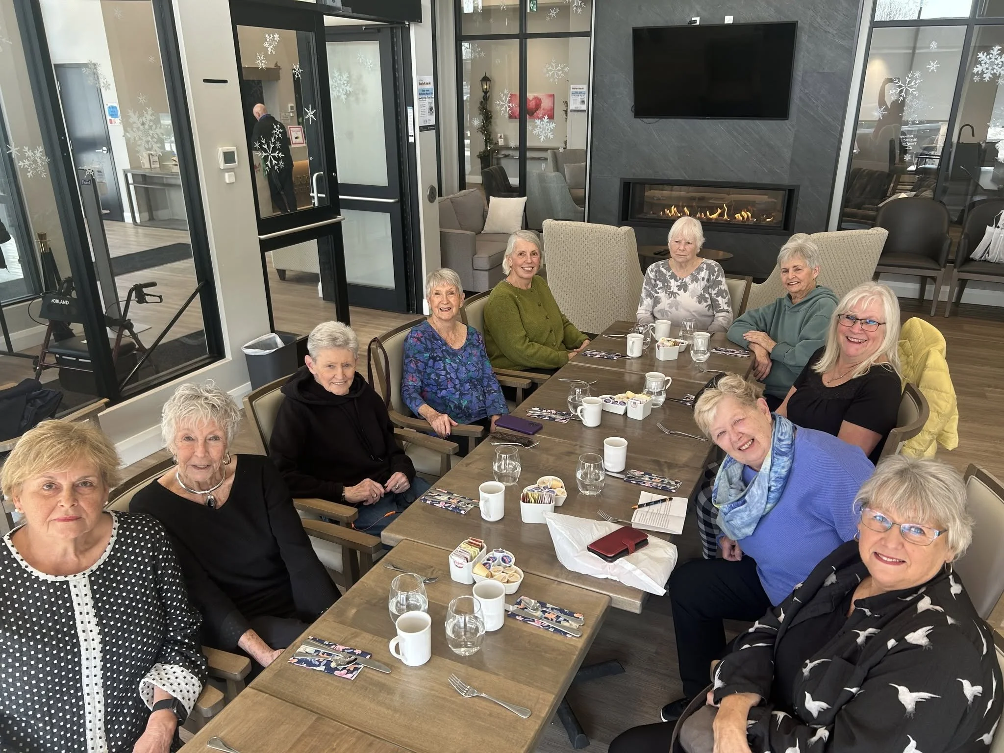 A group of elderly women sitting at a table during a festive gathering in a restaurant or community center, with snowflake decorations on the windows and a fireplace on the wall.