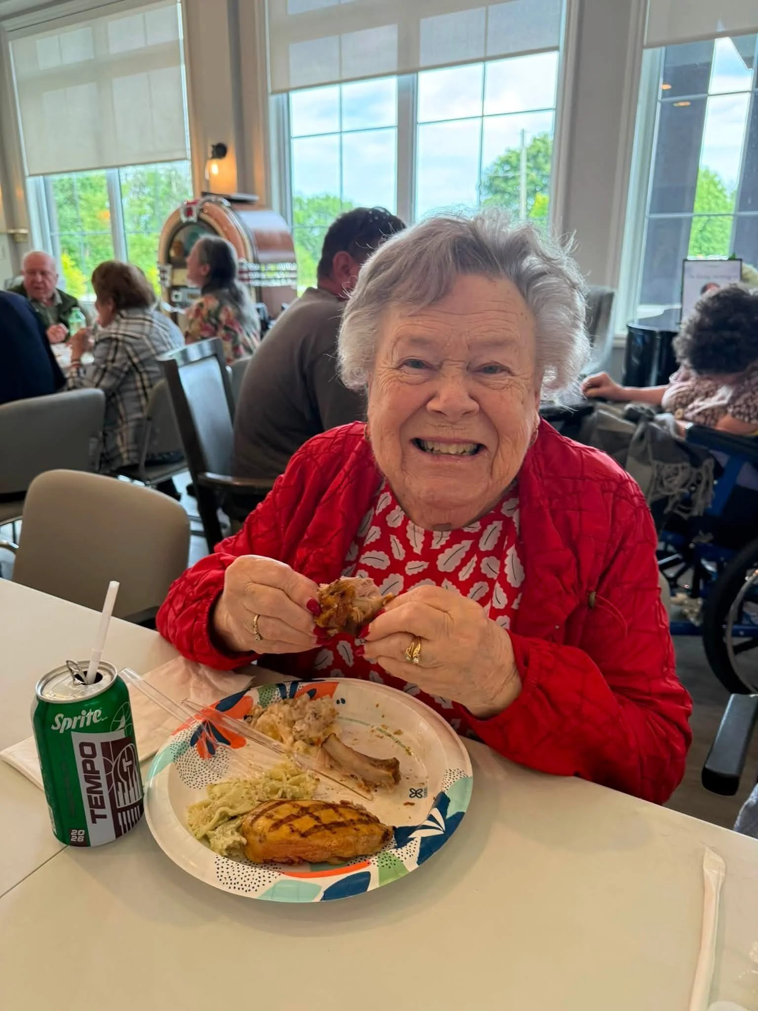 An elderly woman with gray hair smiling at the camera while sitting at a table in a restaurant or cafeteria, holding a piece of food. The table has a disposable plate with food, a green Sprite soda can, and a cup with a straw. In the background, othe
