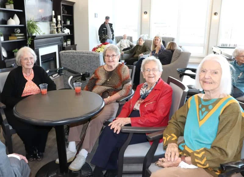 A group of elderly women sitting around a table at a social gathering in a bright room with large windows, with more people in the background.