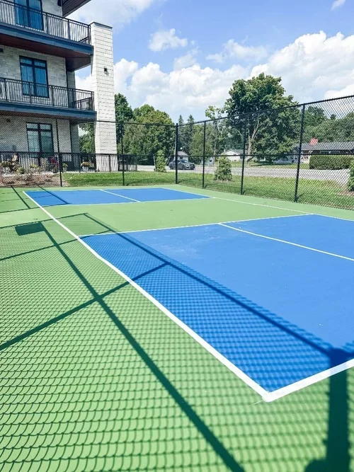 An outdoor tennis court with green and blue surfaces, surrounded by a black fence, with an apartment building and trees in the background.