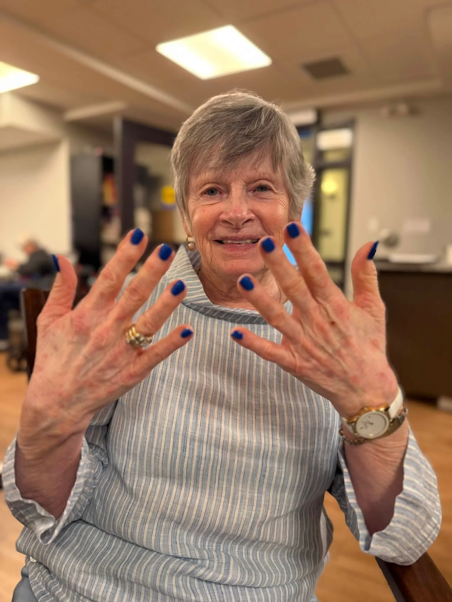 A smiling elderly woman with short gray hair shows her hands with painted blue nails, wearing a striped shirt and jewelry, in a well-lit indoor setting.