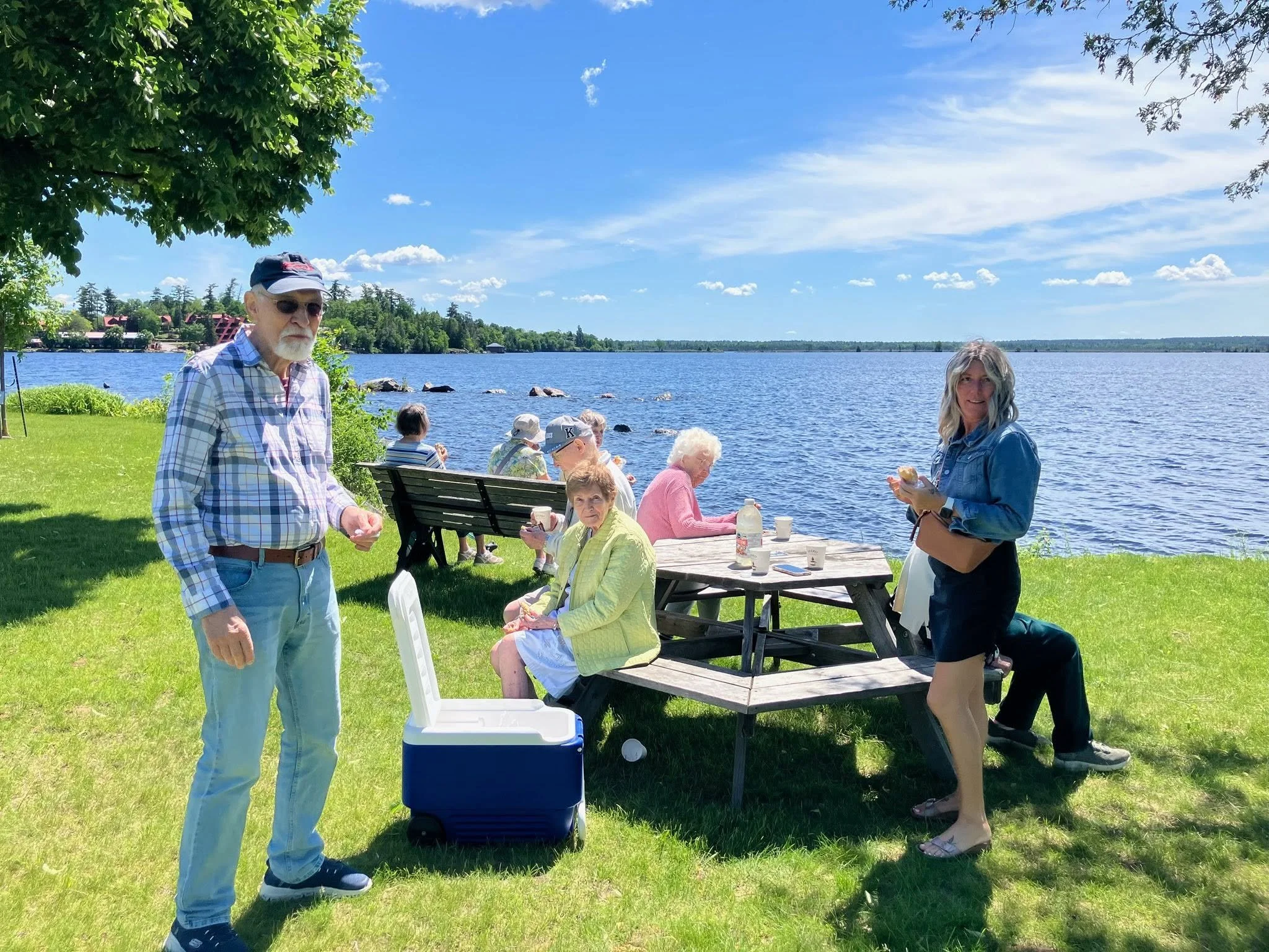 Group of people enjoying a lakeside picnic on a grassy area with trees, a picnic table, and a view of the lake under a partly cloudy sky.