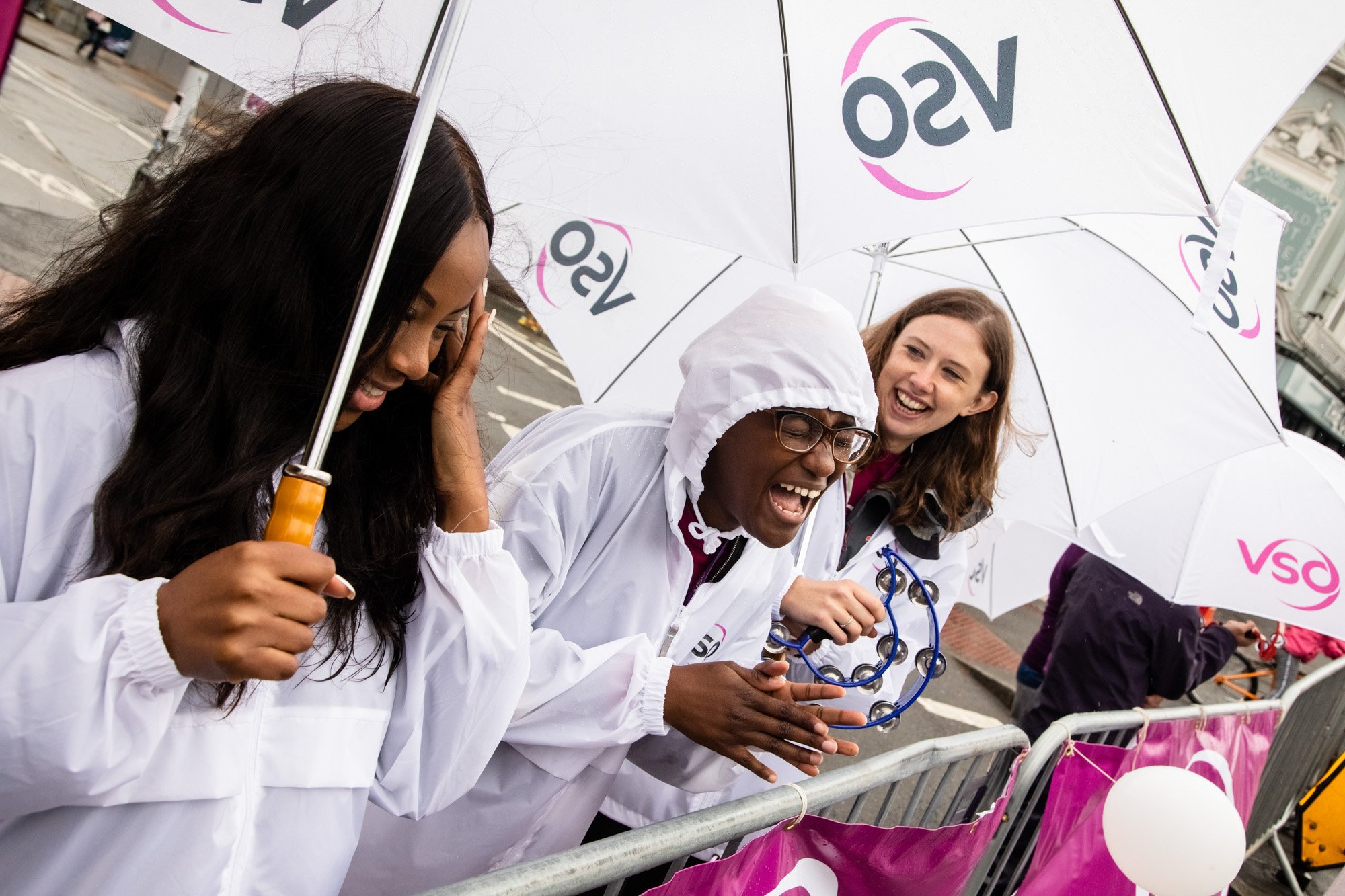 VSO volunteers laughing with tambourines beneath umbrellas as they support RideLondon cyclists.